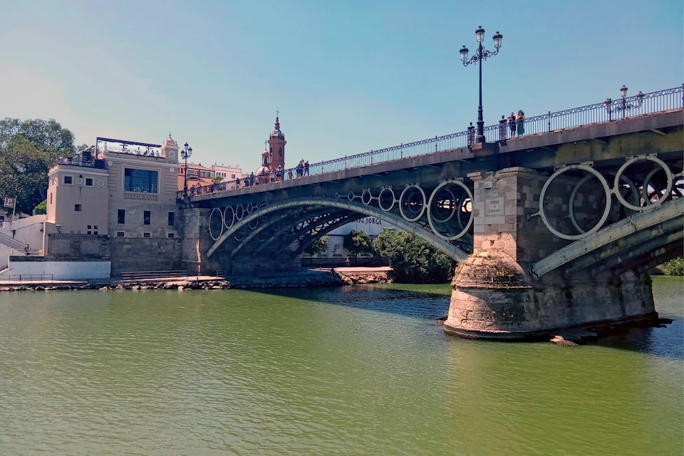 A historic bridge with intricate arches crosses a river, with people walking on it and buildings visible in the background.