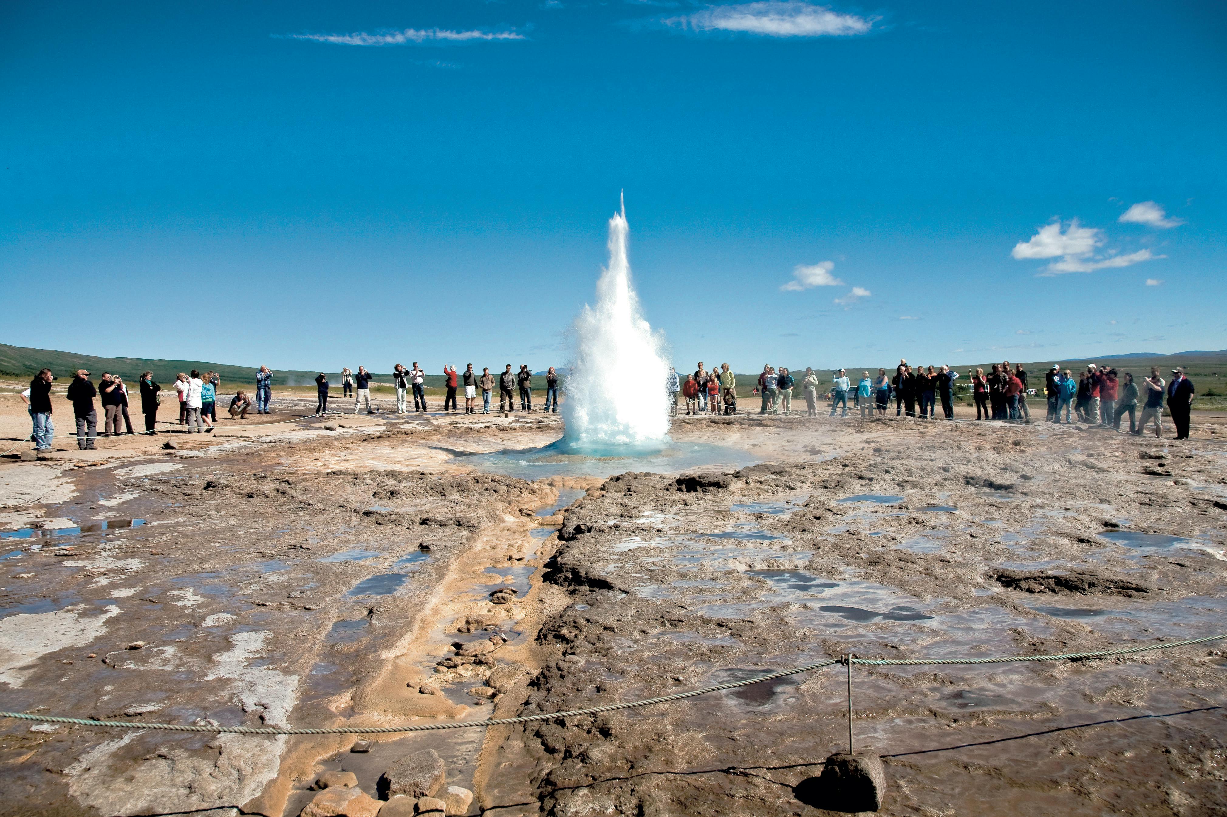 Pessoas que observam a erupção de um géiser sob um céu azul límpido, rodeadas por um solo rochoso e húmido.