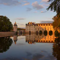 Château avec des ponts en arc au-dessus d'une rivière réfléchissante, des arbres de part et d'autre, et un ciel nuageux éclairé par un coucher de soleil.