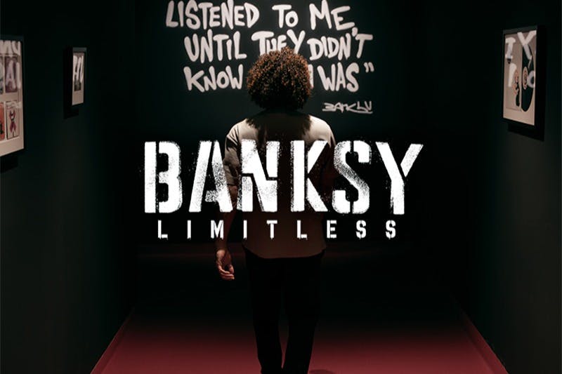 Osoba stojí před graffiti textem, který čte, "POSLOUCHAL MĚ, DOKUD NEVĚDĚL, ŽE JSEM," se slovy "BANKSY NEOMEZENÝ" překryté.