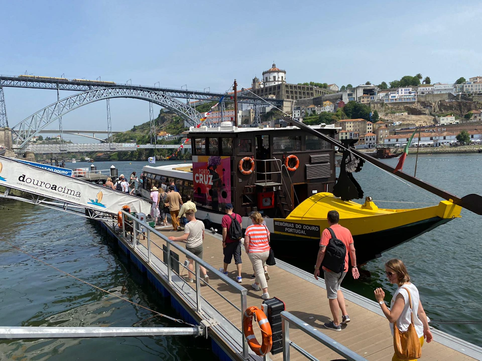 People walking on a dock boarding a boat with a colorful exterior, near a large bridge and hillside buildings in the background.