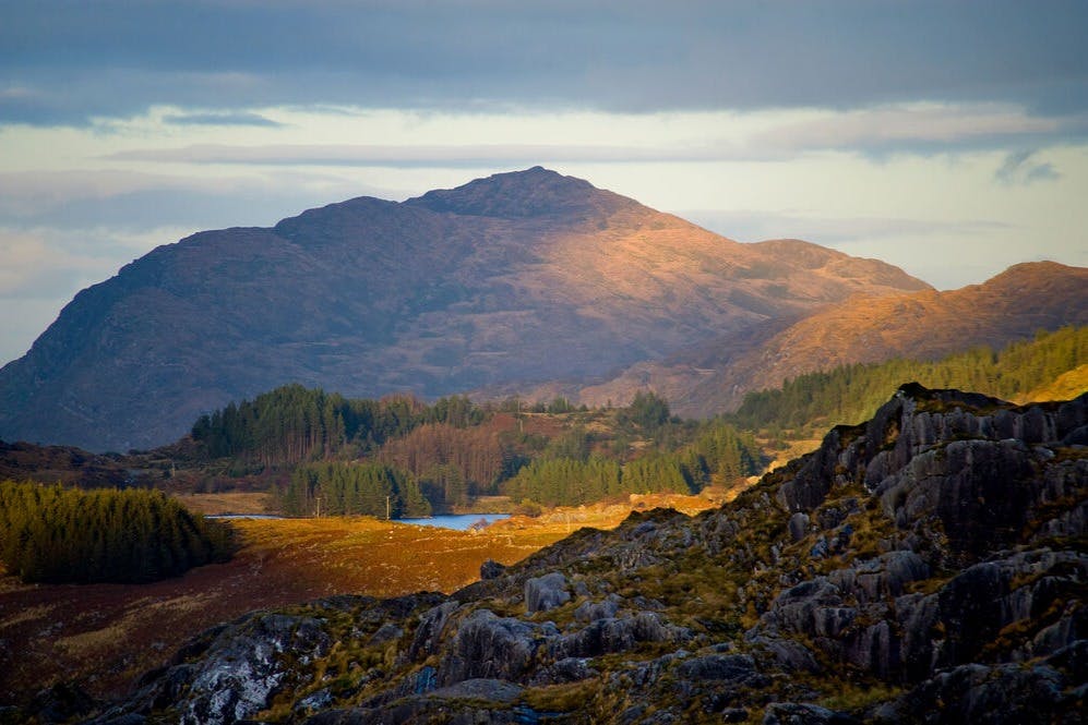 A scenic landscape featuring a mountain lit by sunlight, surrounded by forests and rocky terrain under a cloudy sky.