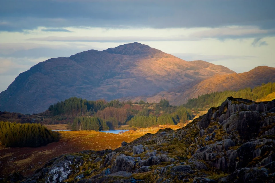A scenic landscape featuring a mountain lit by sunlight, surrounded by forests and rocky terrain under a cloudy sky.