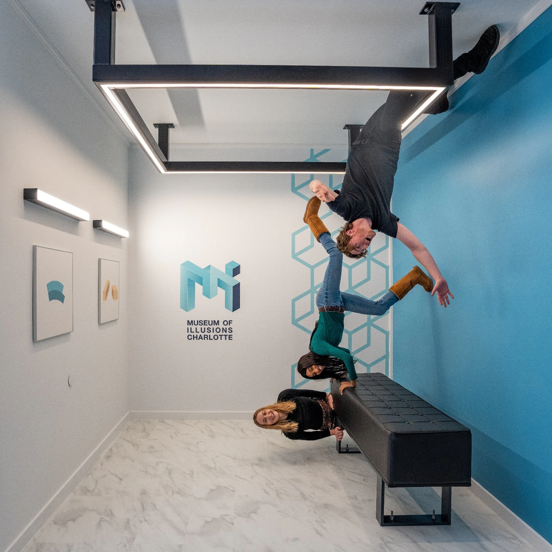 A man appears to hang upside down from the ceiling in a room with optical illusions at the Museum of Illusions in Charlotte. Two women sit on a bench below, creating a playful scene.