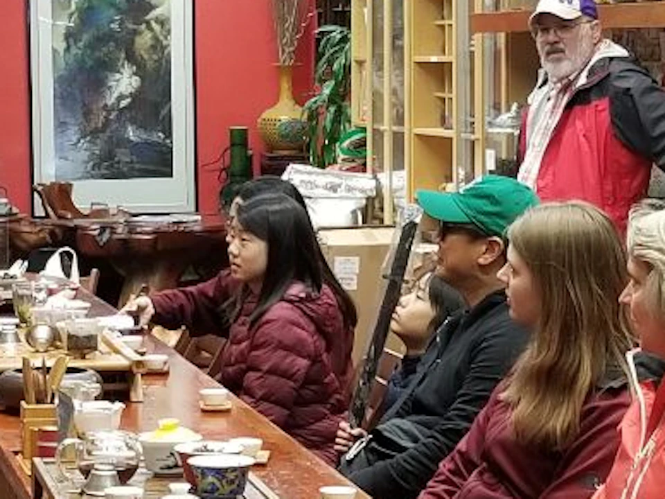 A man stands speaking to a group of seated people in a tea shop, with shelves of tea and teapots visible in the background.