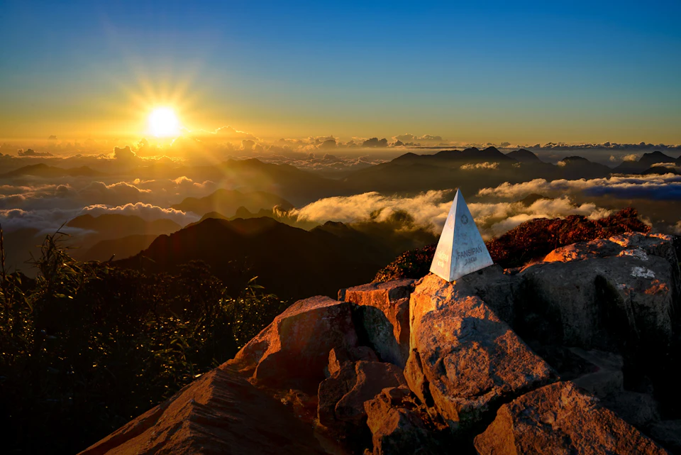 A sunrise view from a mountain peak with a triangular marker surrounded by rocks and clouds below.