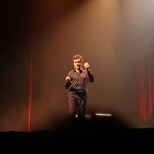 En man i mörk outfit uppträder under scenljus med röda gardiner i bakgrunden.