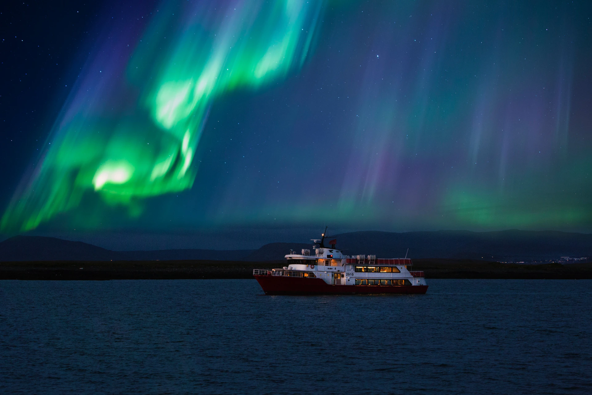 A red and white boat sailing in Faxaflói bay under the northern lights.