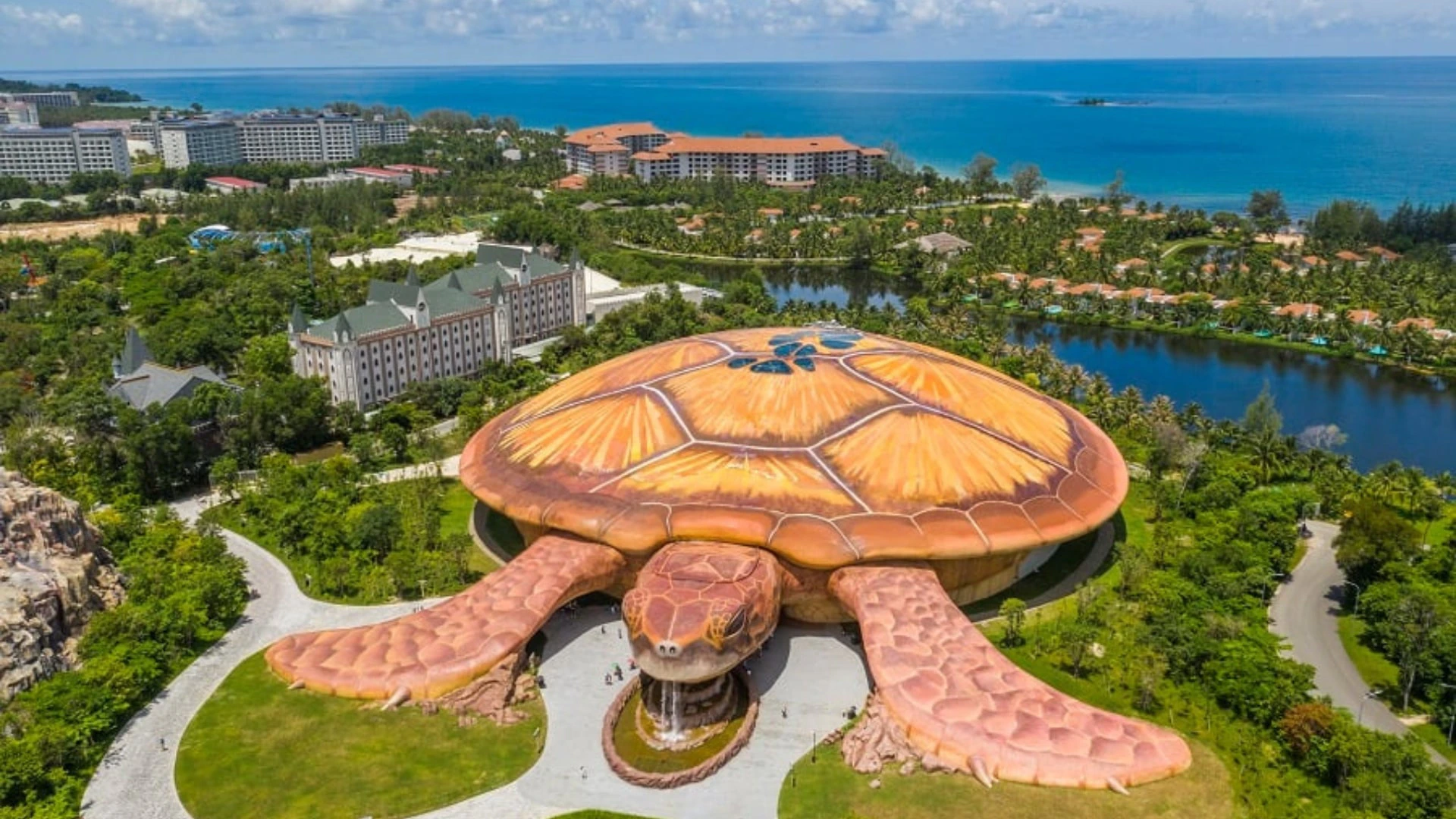 A large turtle-shaped building surrounded by greenery and near a coastal area with resorts in the background.