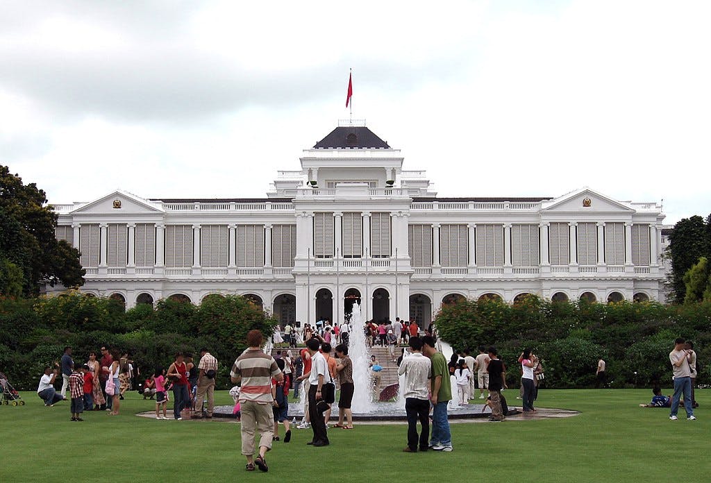 A large white neoclassical building with a central fountain and garden, surrounded by many people walking and gathering.