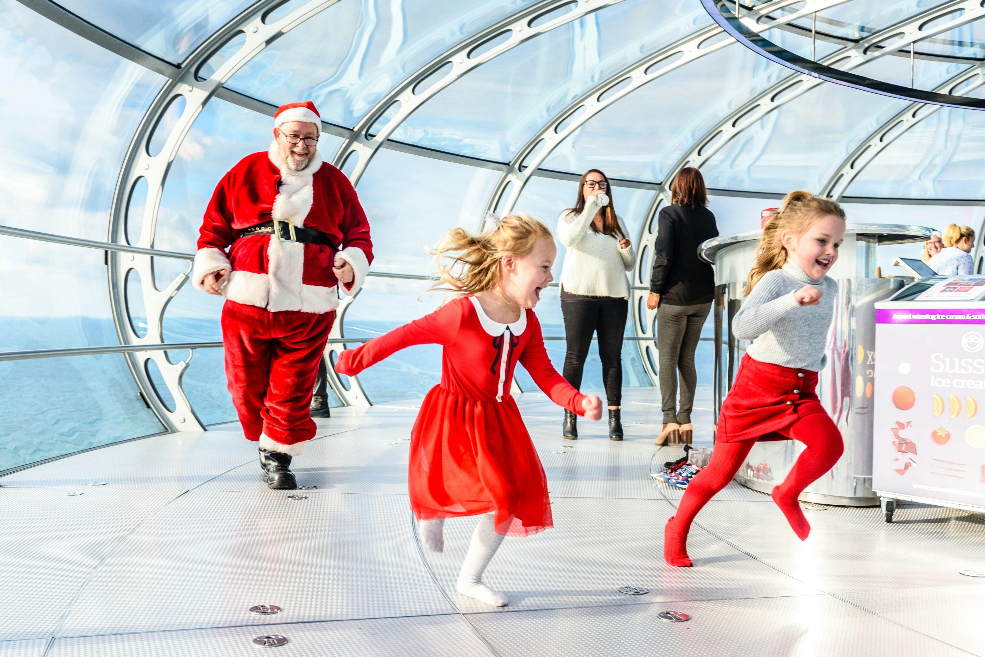 Two girls in red outfits run excitedly in a glass-domed structure. Santa Claus and three adults stand in the background.