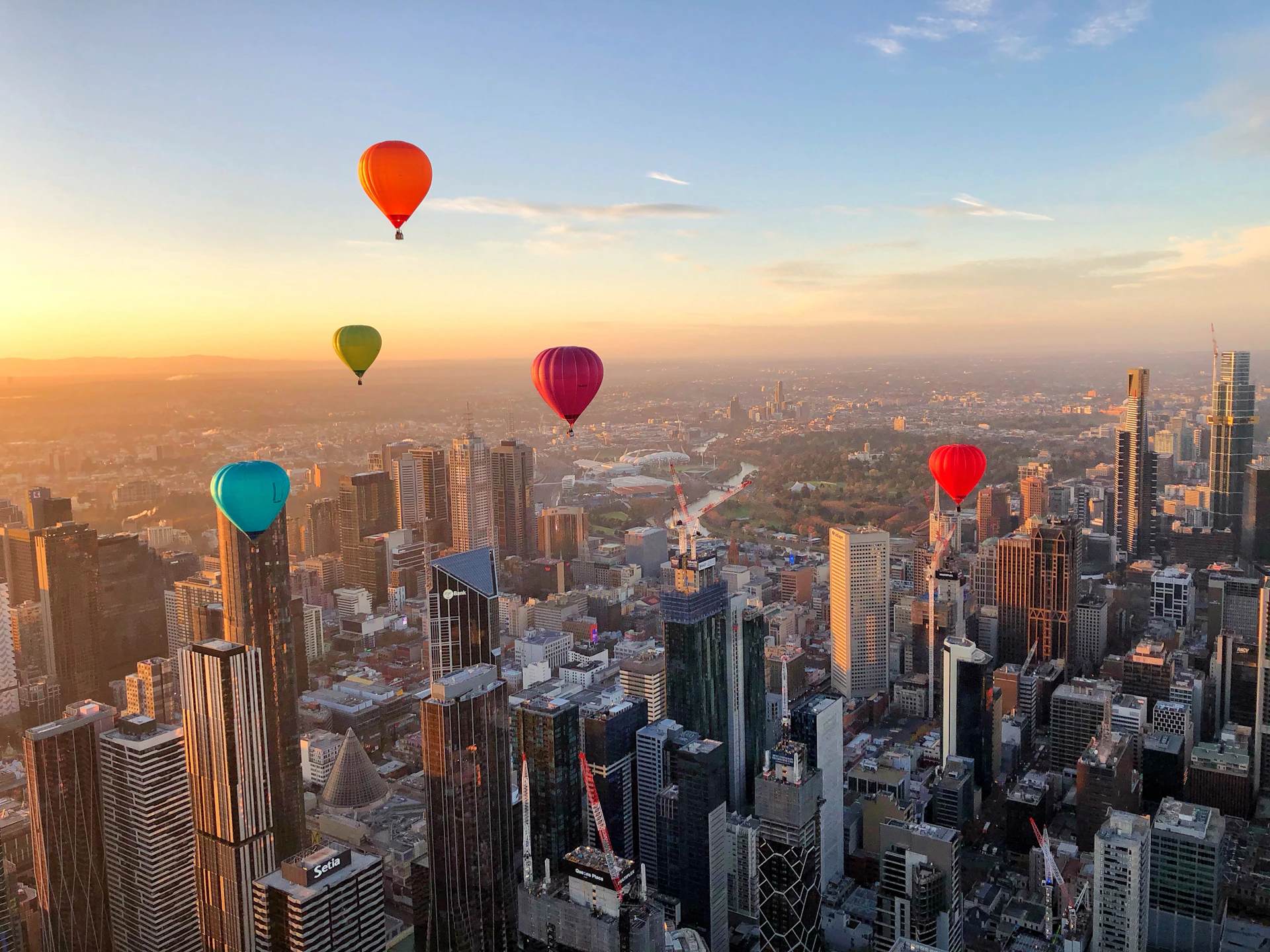 Colorful hot air balloons floating above a city skyline at sunset, with tall buildings and a sprawling urban landscape below.