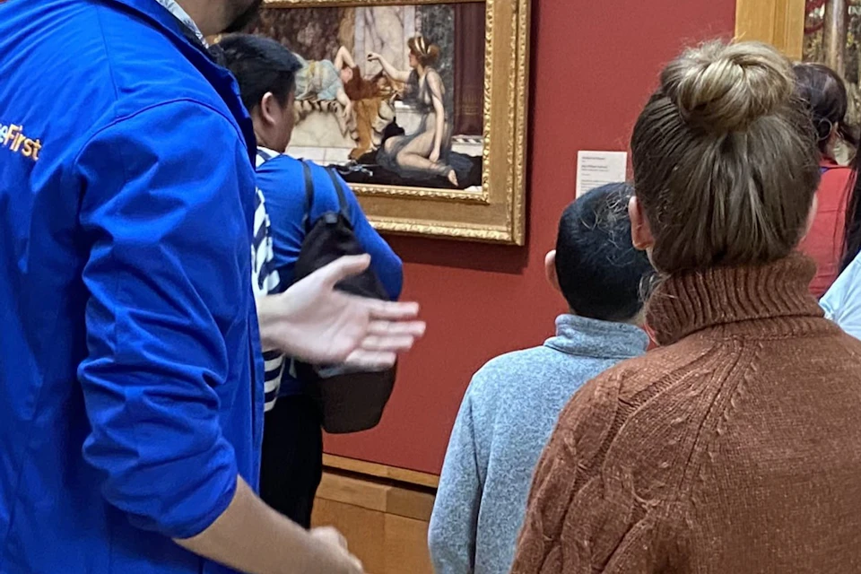 People observing art in a museum room with red walls. A guide wearing a blue jacket is speaking to a group. Busts and paintings are visible.