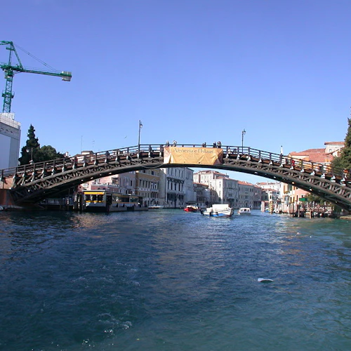 En välvd träbro sträcker sig över en kanal med båtar i Venedig, Italien, under en klarblå himmel. En kran och byggnader är synliga.