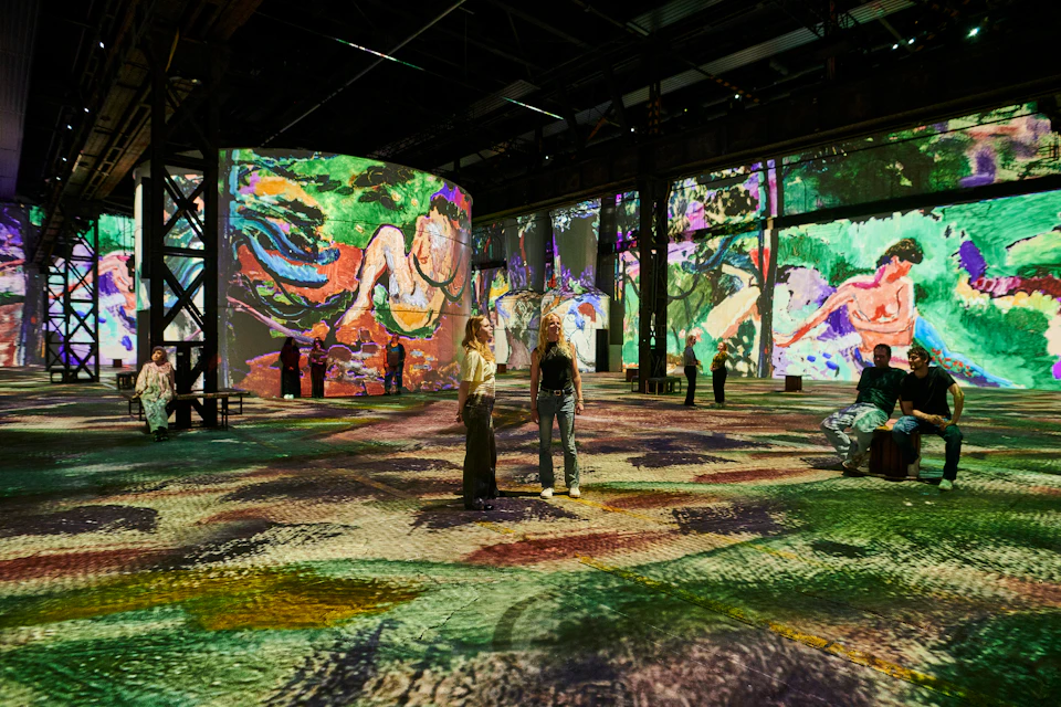 People viewing colorful art projections on walls and floor in a large, dark indoor exhibition space.