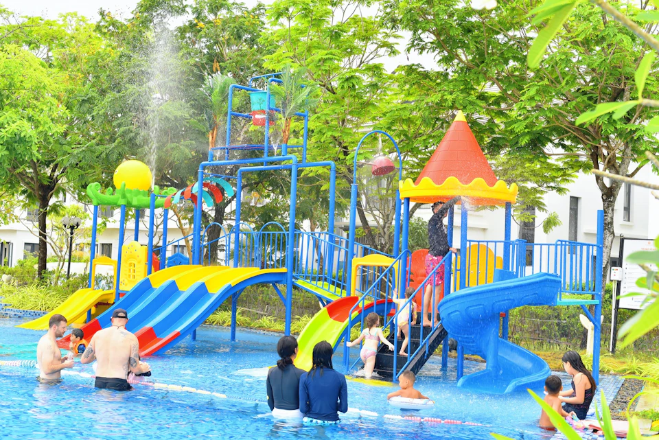 Children and adults enjoying a colorful water play area with slides and sprinklers in an outdoor pool surrounded by trees.