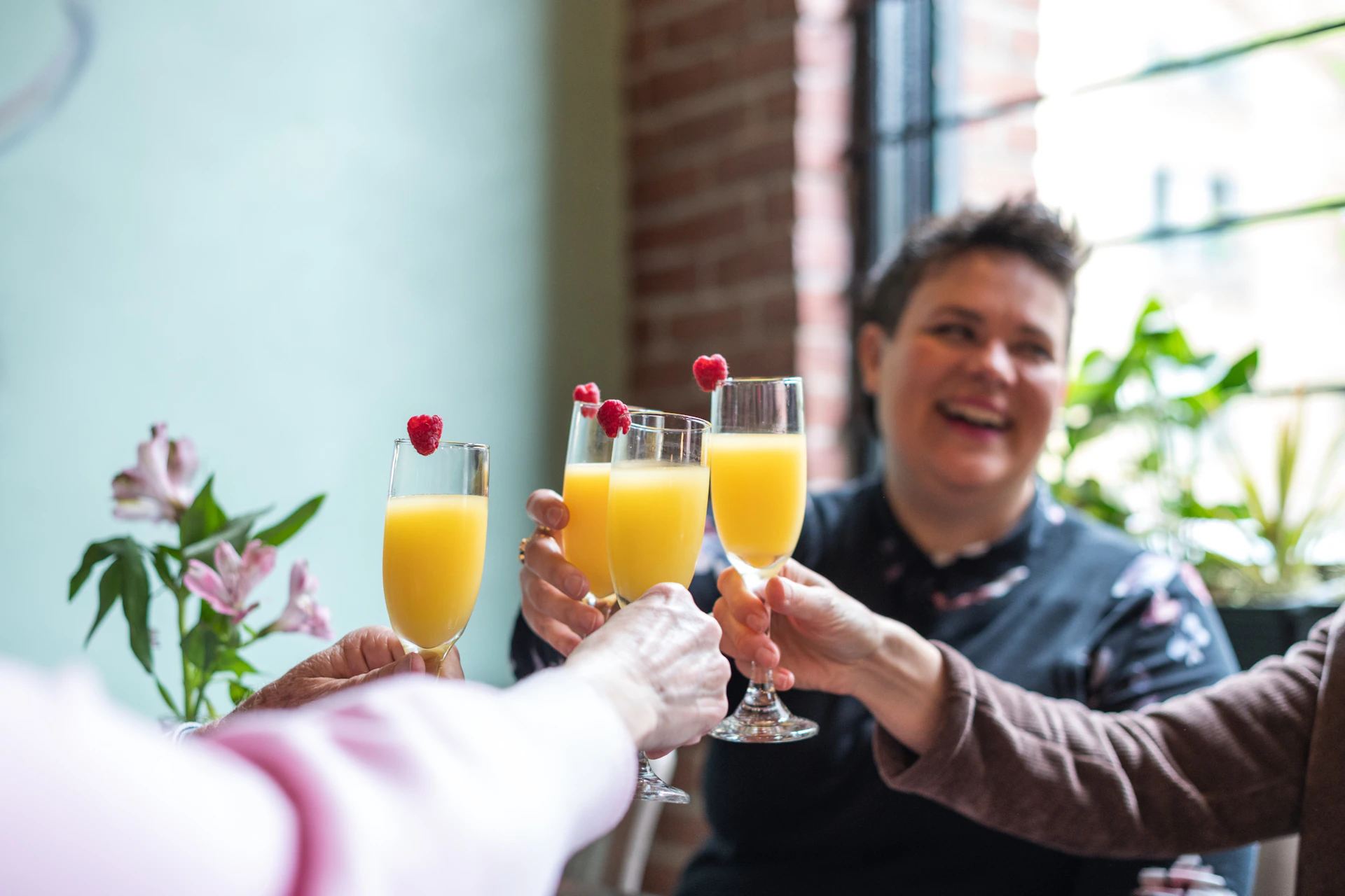 Four people clink glasses of orange drinks with raspberries. A person in the background is smiling, and there are plants near a window.