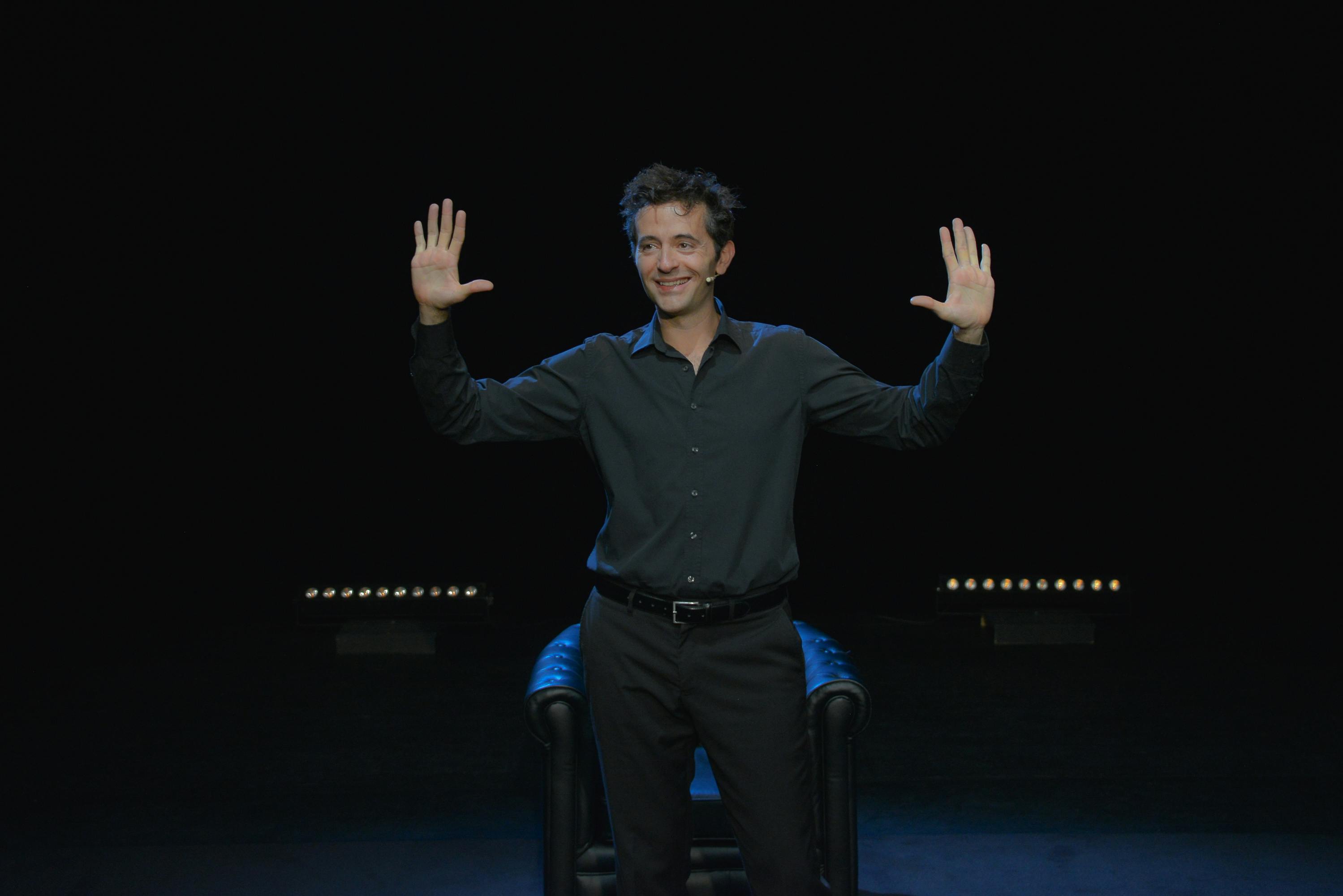 一位身穿深色衬衫的人举起双臂，双手张开，微笑着站在黑色背景和皮椅前。