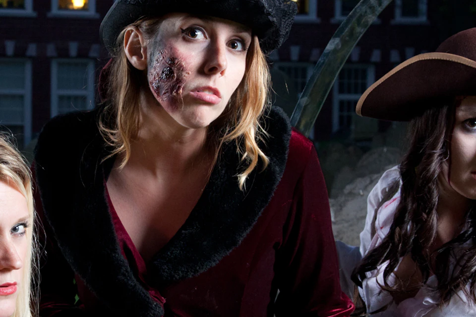 Three people in costume, with eerie makeup, standing behind tombstones in a cemetery at night. Lantern in the foreground.