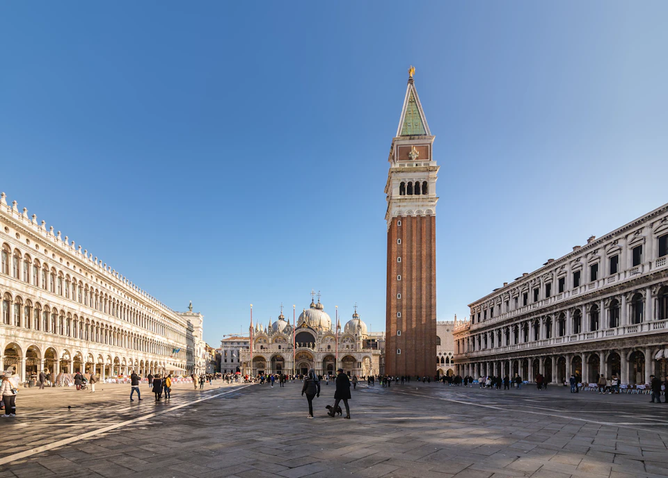 Venice's St. Mark's Square with the Campanile tower, St. Mark's Basilica, and surrounding historical buildings under a clear blue sky.