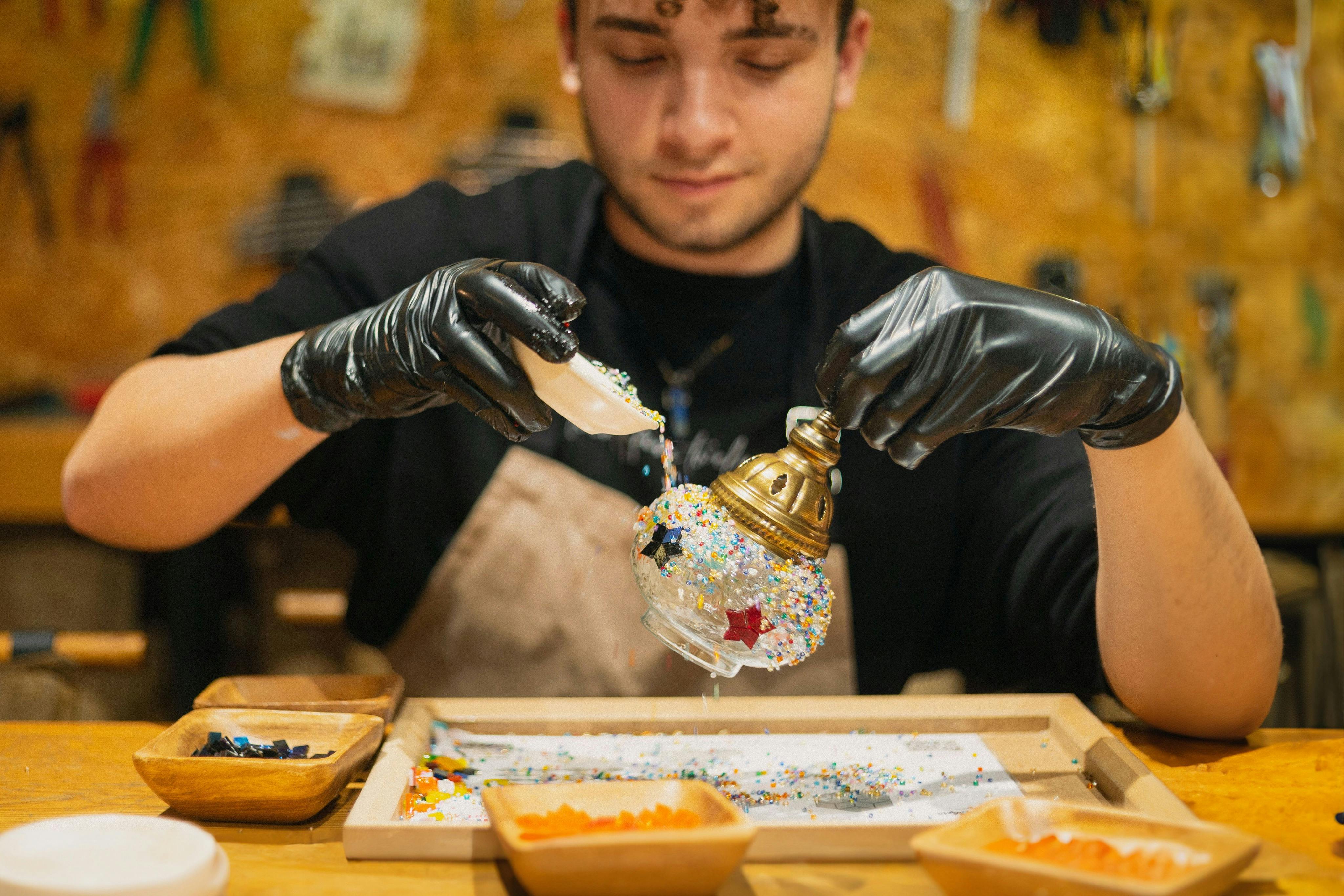 The participant in black gloves crafting a Turkish mosaic lamp, pouring colorful stones onto a glass sphere in a workshop set