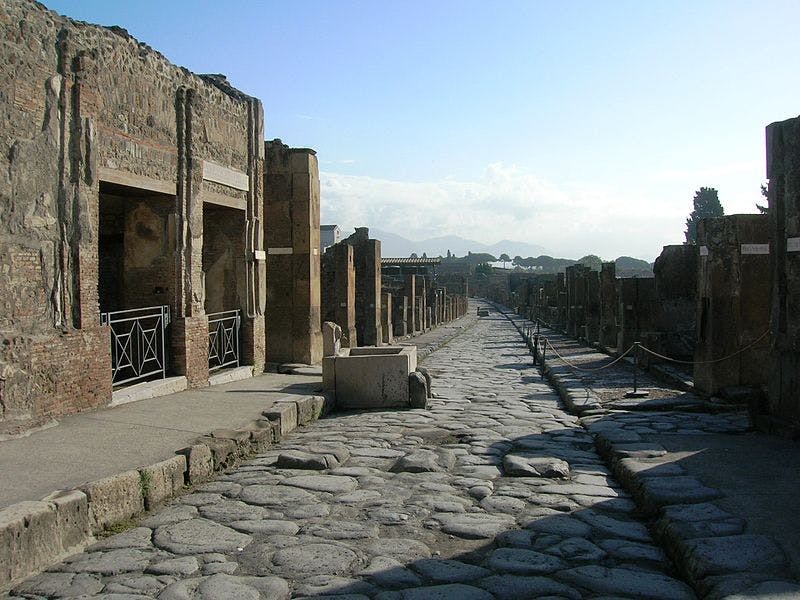 Ancient stone-paved street with ruins of buildings on both sides, under a clear sky with distant mountains.