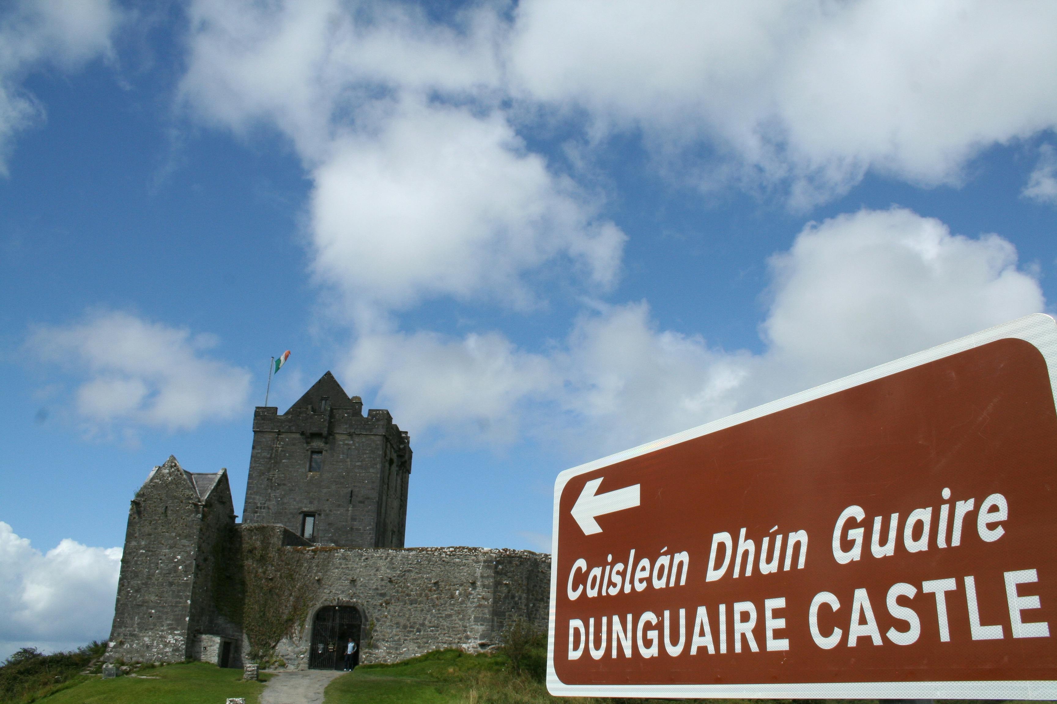 Stone castle with an Irish flag on a tower under a blue sky with clouds. Brown directional sign reads "Dunguaire Castle" in two languages.