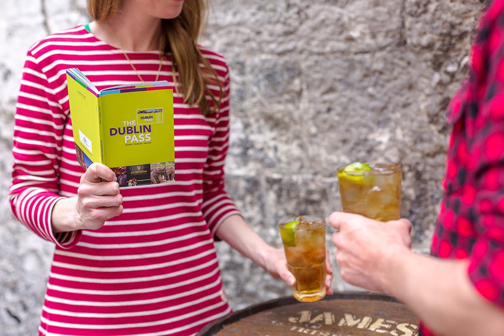 Woman in a red striped shirt holding a &#34;Dublin Pass&#34; book, while two people hold drinks with lime slices.