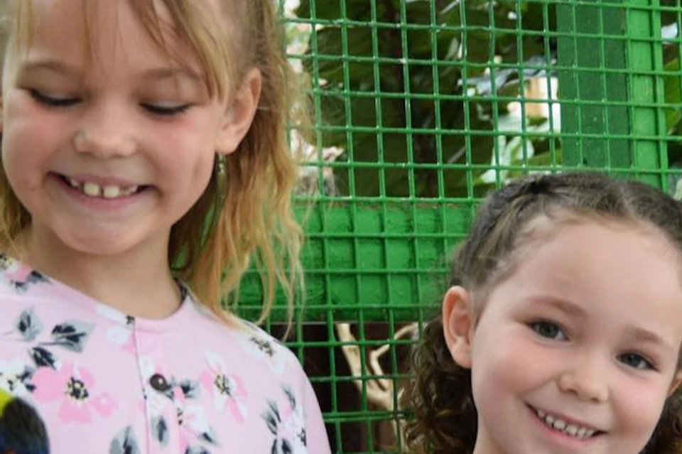 Two young girls smile while holding colorful parrots inside a green mesh enclosure with foliage in the background.