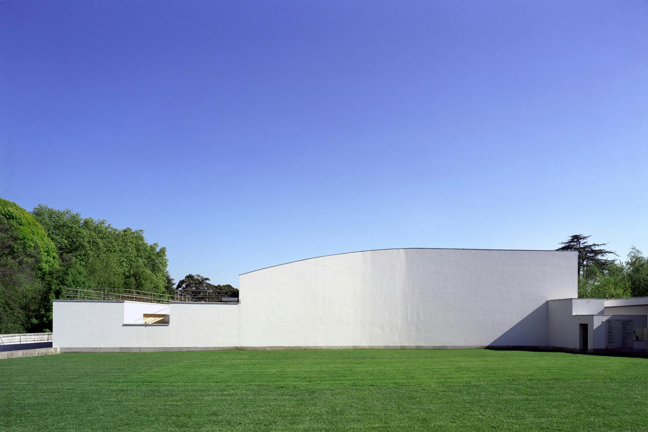 Flat grass field in front of a white, curved-wall building with a clear blue sky above. Trees are seen on the left side.