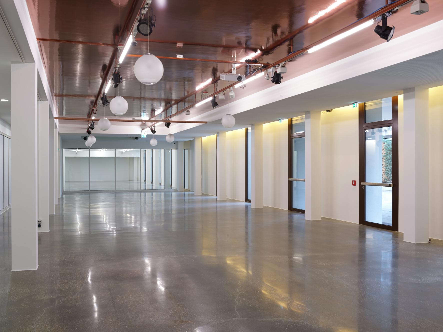 Ένας μεγάλος, άδειος εσωτερικός χώρος με ανακλαστικά δάπεδα, φωτιστικά οροφής και μεγάλα παράθυρα κατά μήκος της μίας πλευράς.