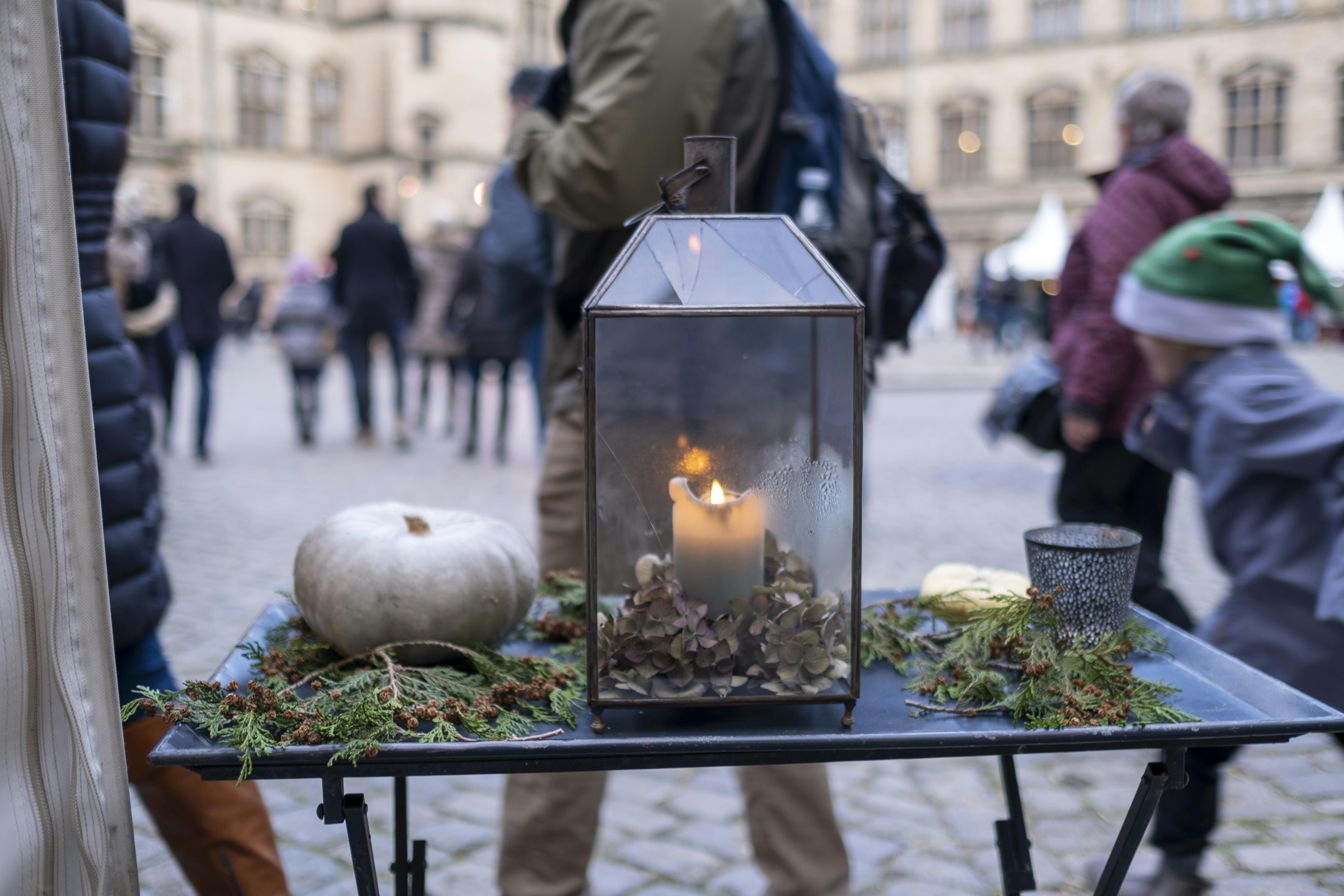 A glass lantern with a lit candle, a white pumpkin, and greenery on a table in an outdoor plaza with people in the background.