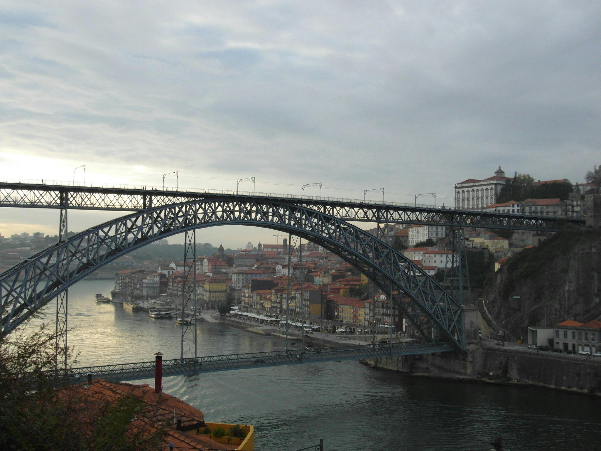 En dubbeldäckad metallbro sträcker sig över en flod, med en stadsbild av byggnader med röda tak i bakgrunden under en molnig himmel.