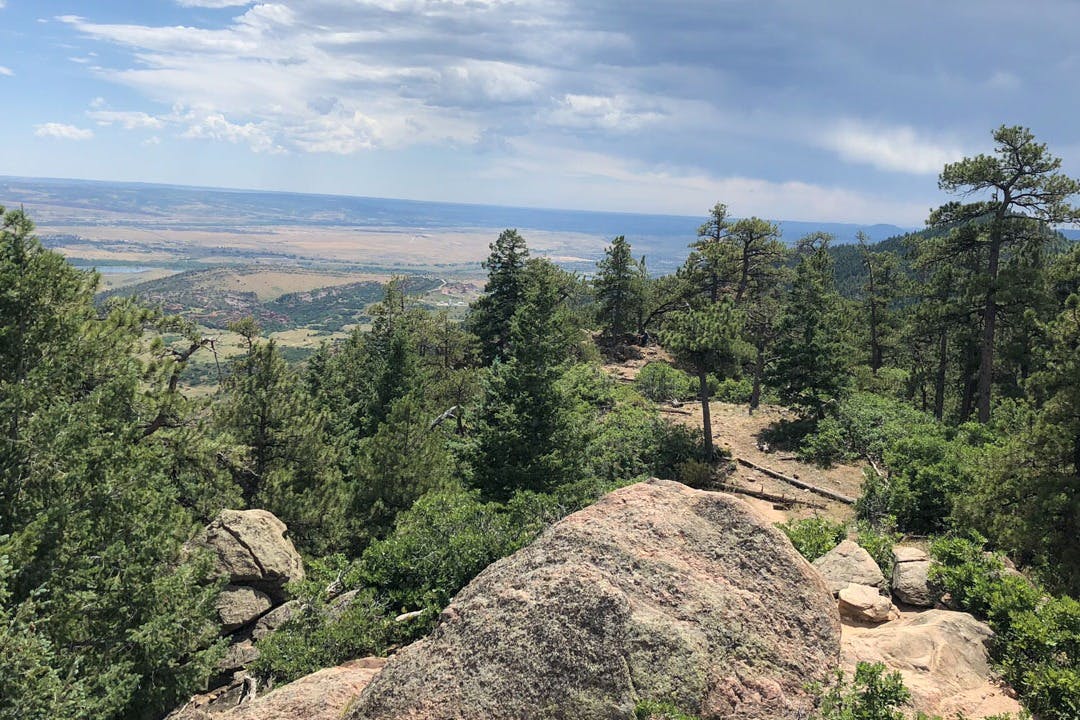 Paisatge de muntanya amb un primer pla rocós, densos arbres verds i un altiplà llunyà sota un cel parcialment ennuvolat.