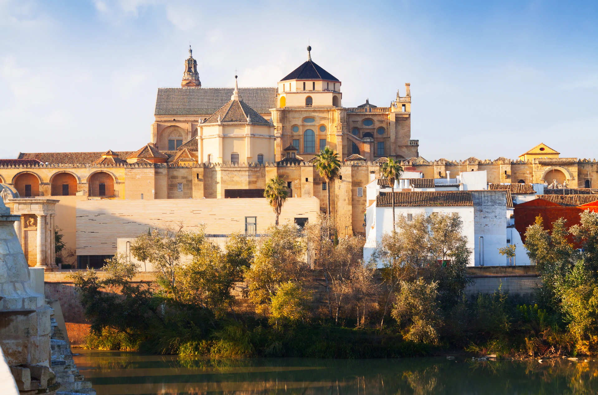 Historic building with domed and spired roofs behind a row of trees and a river, under a clear blue sky.