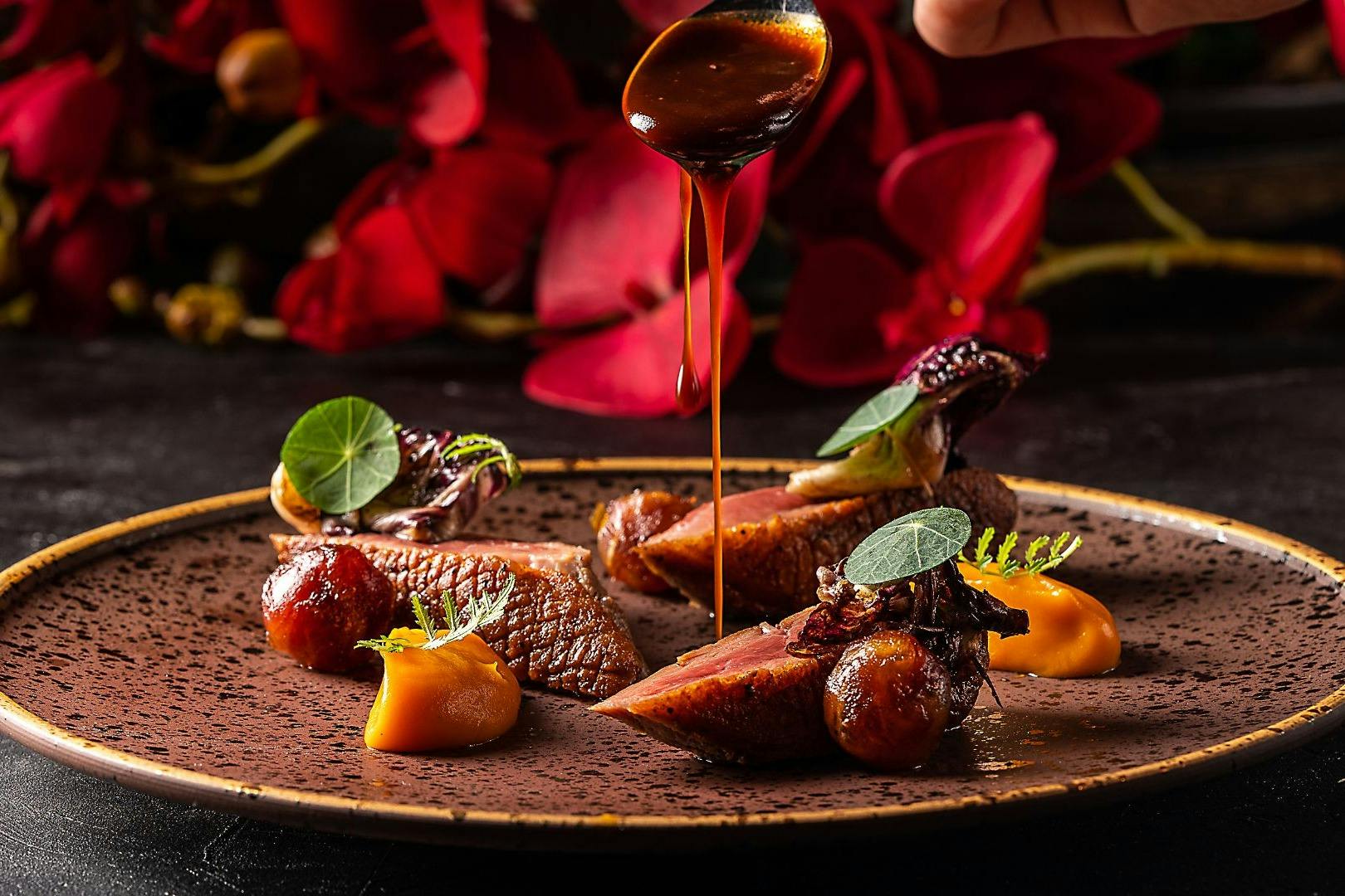 A gourmet dish with seared meat, roasted vegetables, sauces, and garnish, with a spoon drizzling sauce and bright red flowers in the background.