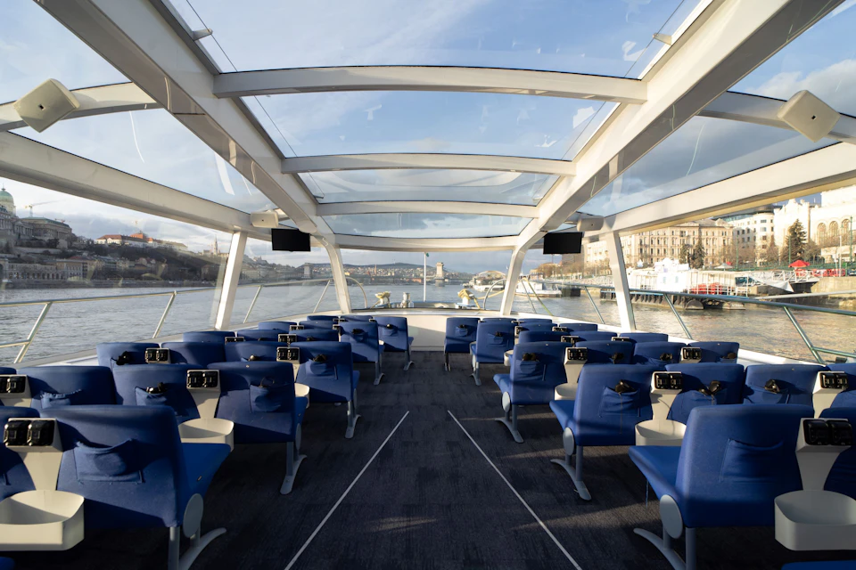 Interior of a modern boat with rows of blue seats, large windows, and a glass roof, overlooking a river and cityscape.