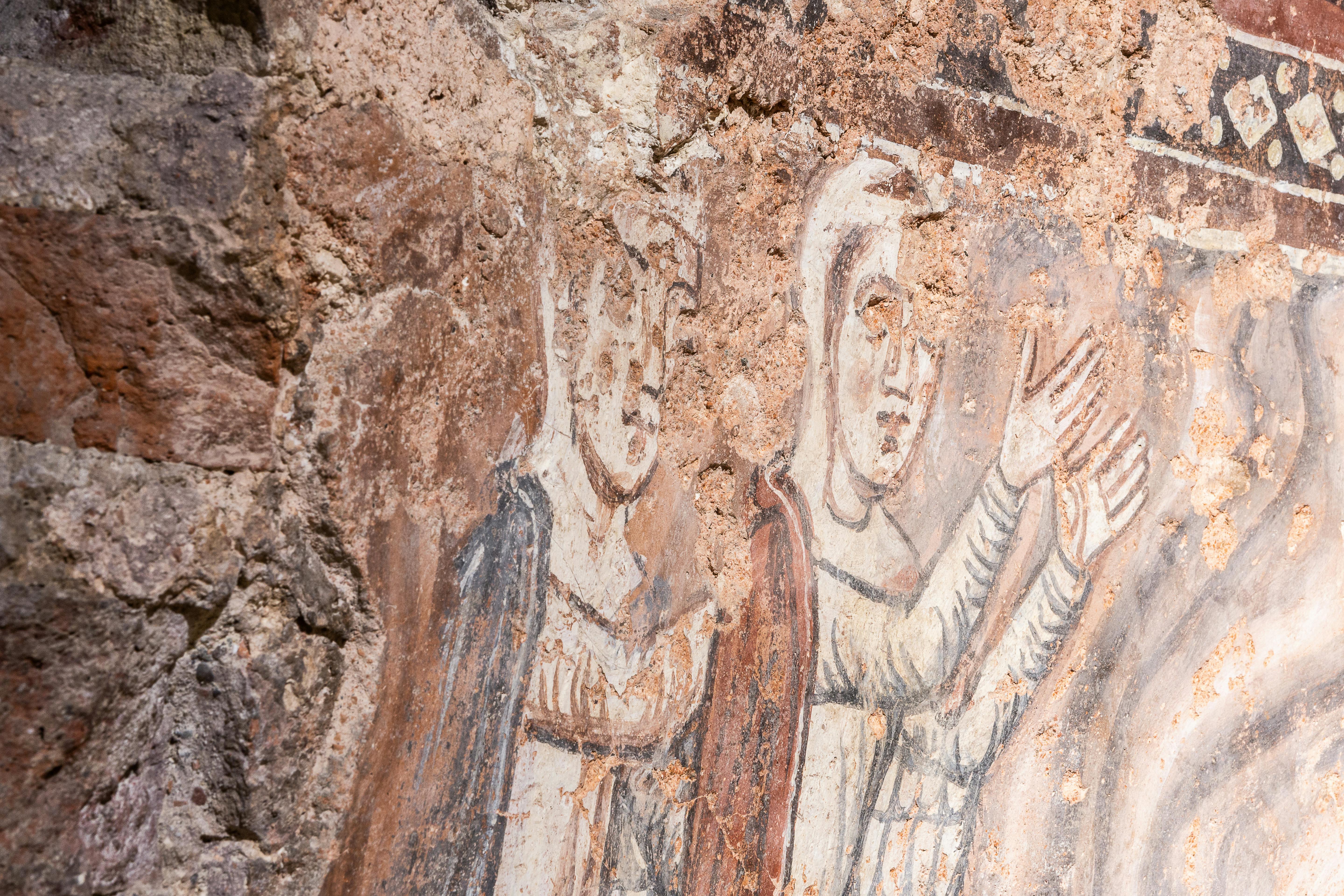 Two faded medieval frescoes on a rough stone wall depicting figures with raised hands, wearing robes and headdresses.