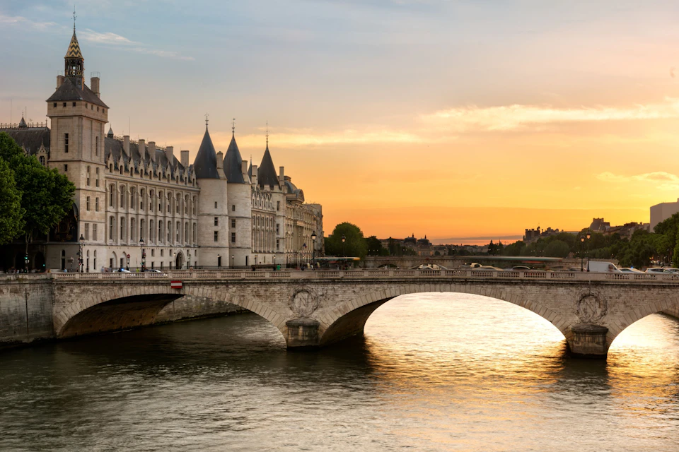 A historic building with towers by a river, adjacent to a stone bridge, under a vibrant sunset sky.