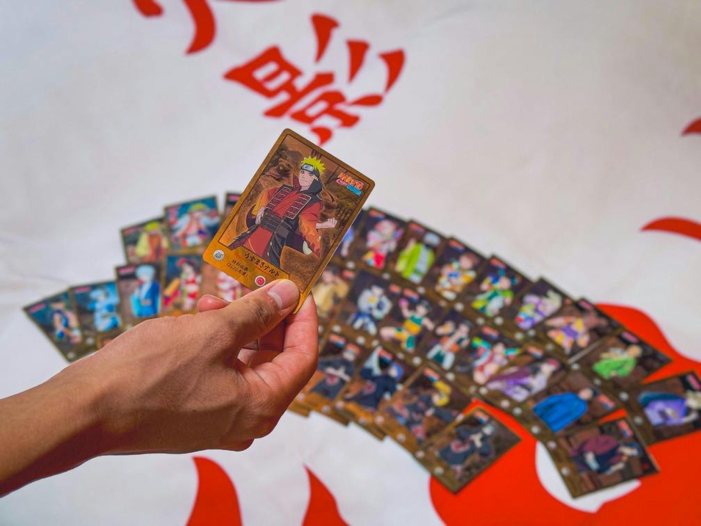A hand holding a trading card featuring an anime character, with more cards displayed on a surface below.