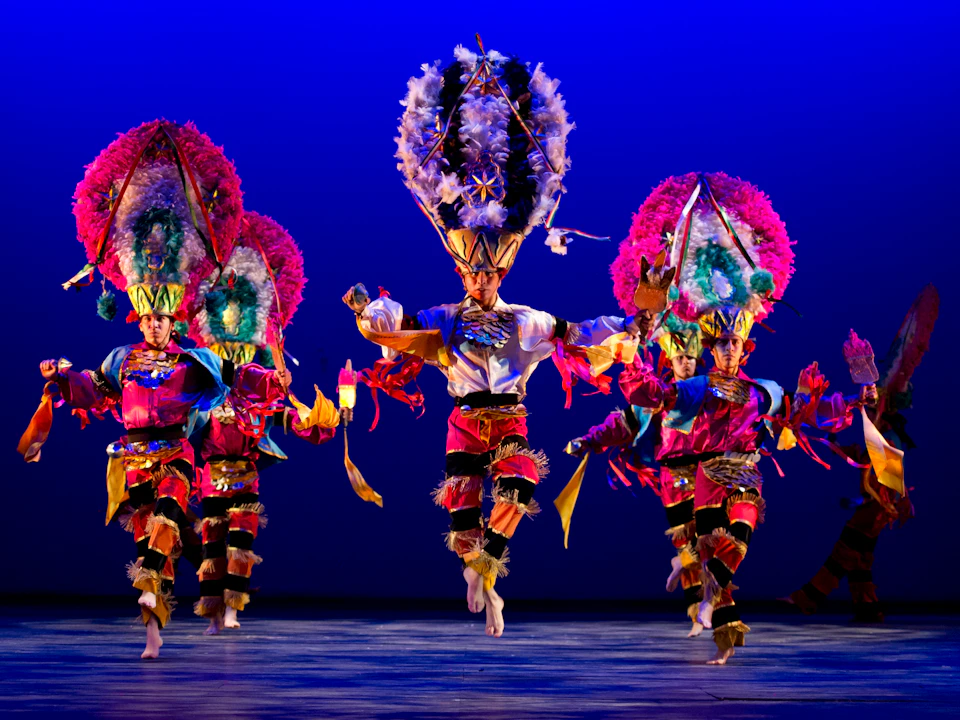 Performers in vibrant, traditional costumes with feathered headdresses dance energetically on stage against a blue background.