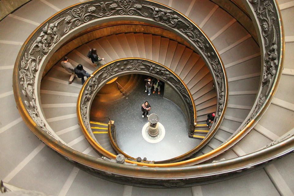 People walking on a spiral staircase with ornate railings, seen from above; a central column at the bottom.