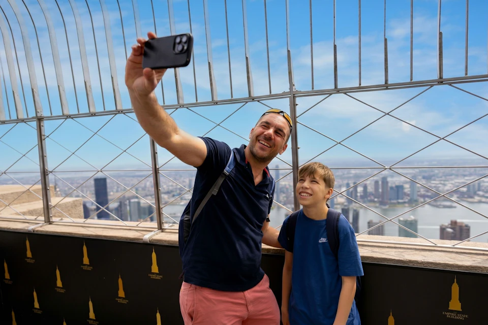 A man and a boy taking a selfie on an outdoor observation deck with a cityscape visible through metal fencing.