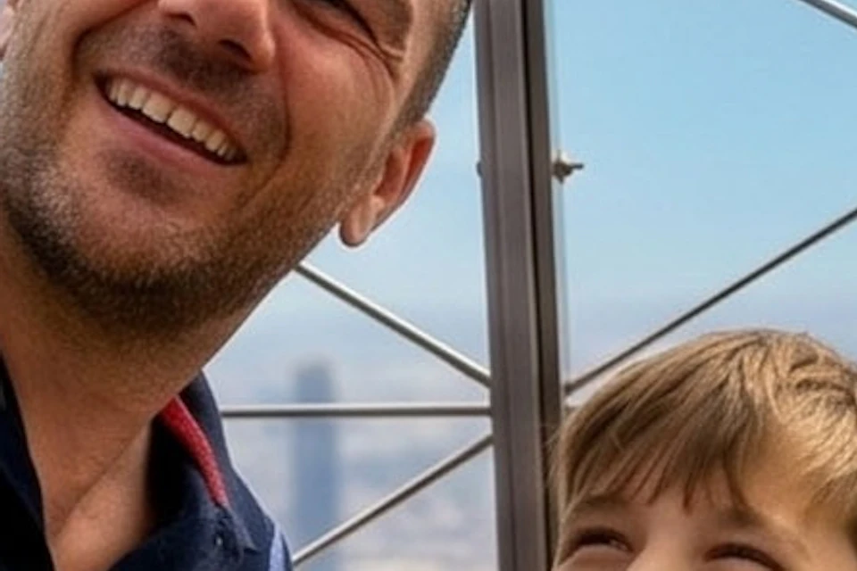 A man and a boy taking a selfie on an outdoor observation deck with a cityscape visible through metal fencing.