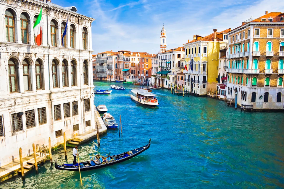 Venice canal with gondolas, boats, colorful buildings, and a tower under a clear blue sky.