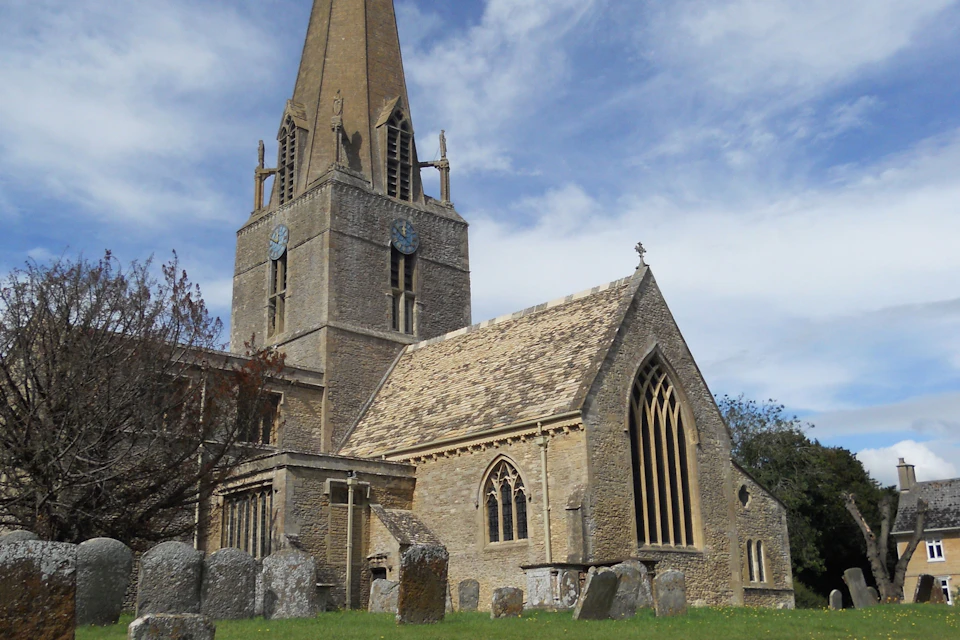 A stone church with a tall spire and clock faces on its tower, surrounded by a graveyard under a partly cloudy sky.