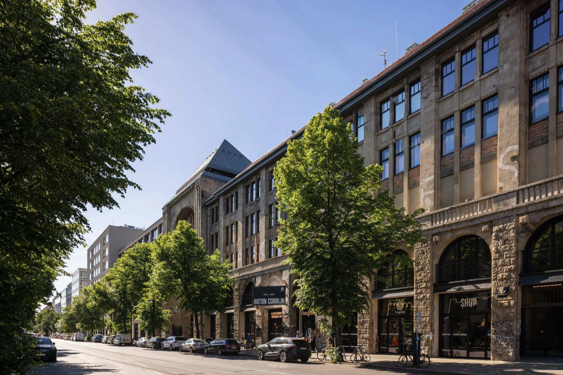 A city street with a row of trees in front of a historic building featuring large windows and arches under a clear blue sky.