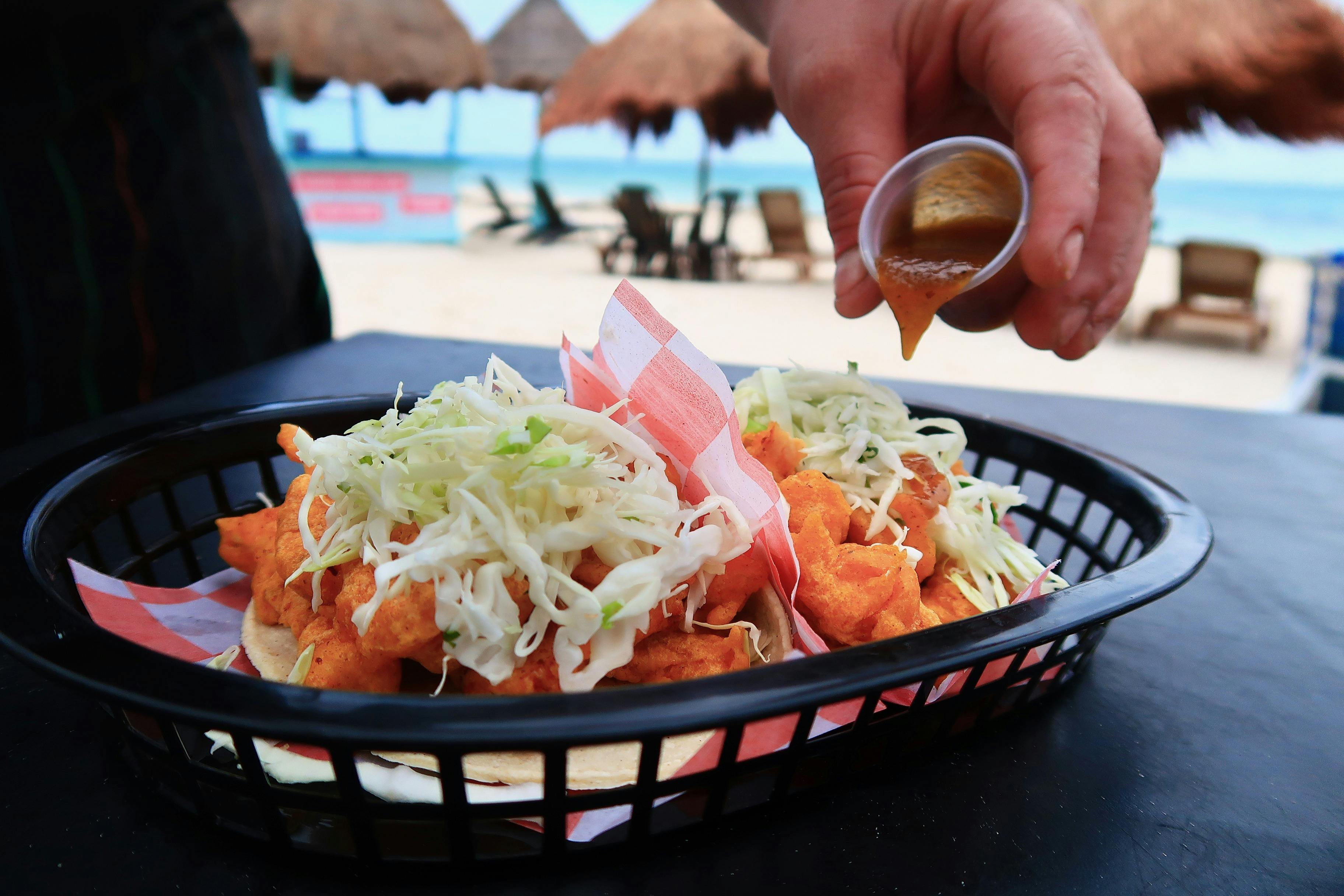 Tacos mit zerkleinertem Kohl in einem Korb, mit einer Hand, die die Soße aus einem Becher gießt, in einer Strandumgebung.