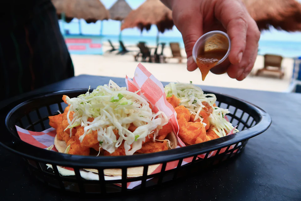 Tacos topped with shredded cabbage in a basket, with a hand pouring sauce from a cup, on a beachside setting.