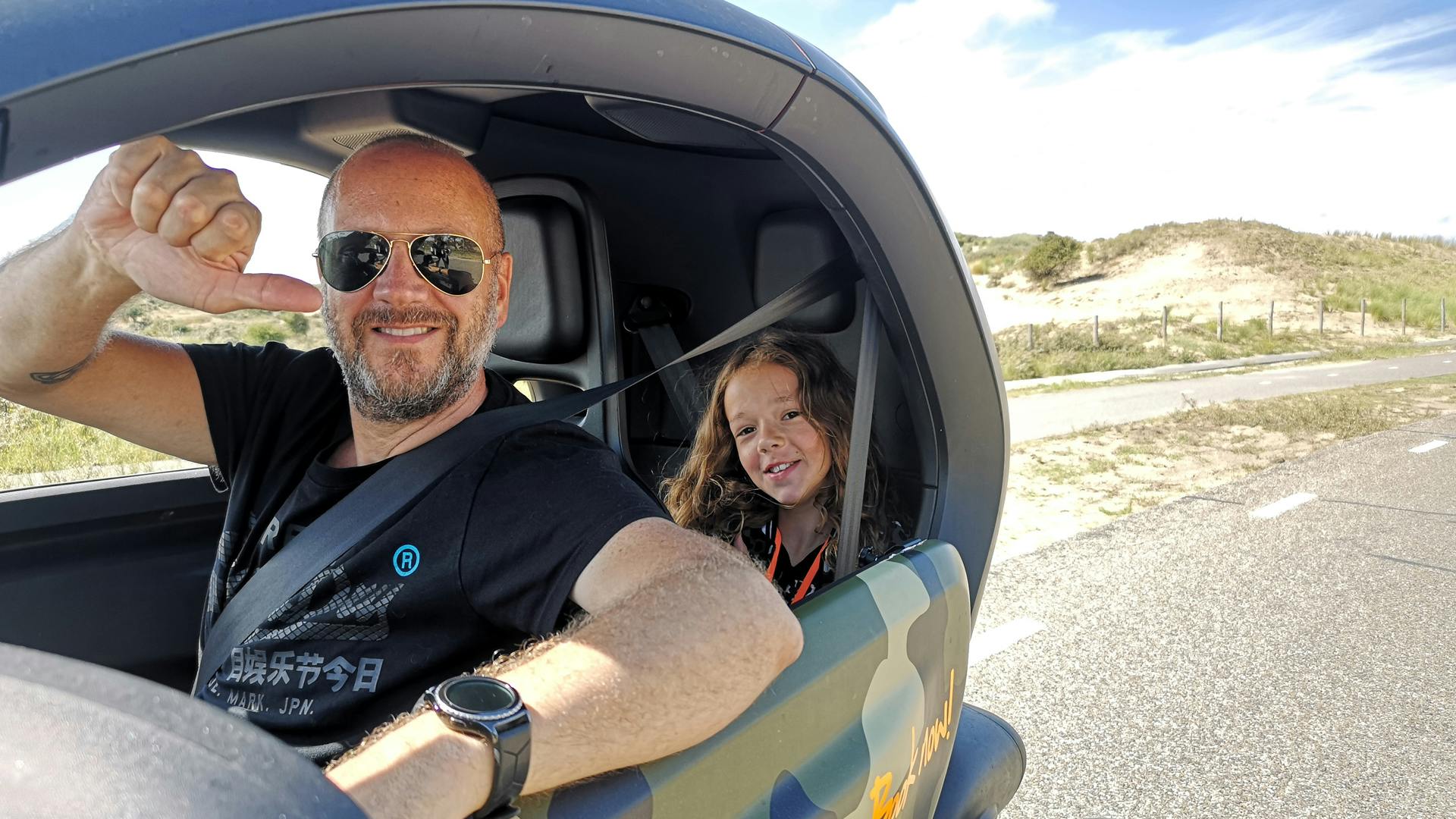 Smilende mand i solbriller og barn med langt hår i en lille bil. Manden giver en tommelfinger op; begge bruger sikkerhedsseler.